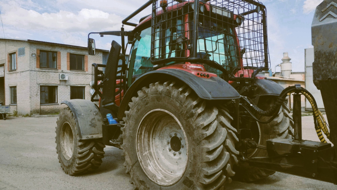 Sarkans traktors pieāķēts pie piekabes. Traktora kabīnei apkārt piestiprināts papildu drošības režģis