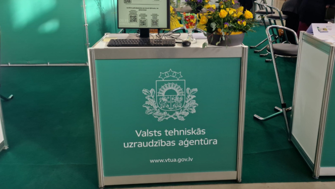 Valsts tehniskās uzraudzības aģentūras stends izstādē biznesa centrā Rāmava. Galds ar Ģerboni uz zaļa fona, uz kura novietots dators e-pakalpojumu saņemšanai