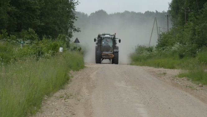 Tumšs traktors brauc pa zemes ceļu, veidojot pēc sevis lielu putekļu mākoni