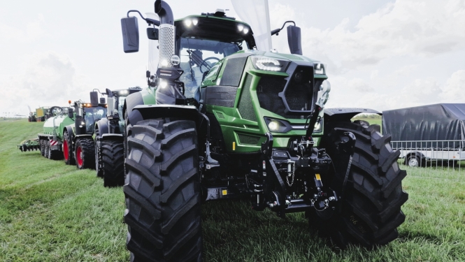 Traktoru kolonna pļavā, priekšā zaļš traktors ar lielām, melnām riepām