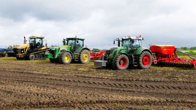 Trīs krāsaini traktori uz lauka ar piekabinātiem dažādiem agregātiem