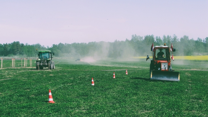 Divi krāsaini traktori brauc pa lauku. Sarkanais traktors brauc pretī, kamēr zaļais prom. Fonā meža ainava