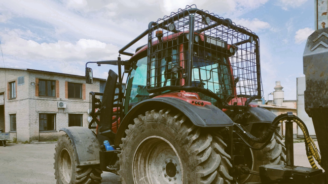 Sarkans traktors pieāķēts pie piekabes. Traktora kabīnei apkārt piestiprināts papildu drošības režģis