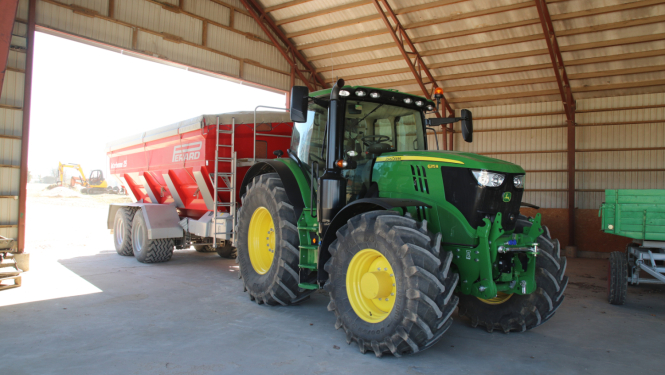 Zaļš traktors ar dzelteniem riteņiem un sarkanu piekabi iebraucis noliktavā. Attēls no traktora priekšas, skatā no noliktavas vidus