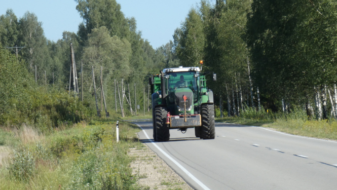 Zaļš traktors ar piekabi brauc pa asfaltētu ceļu. Fonā mežs