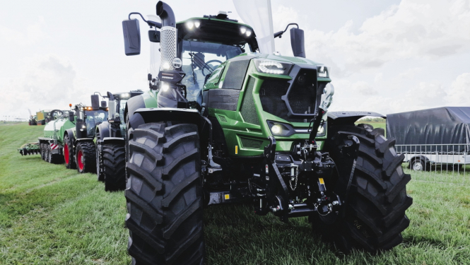 Traktoru kolonna pļavā, priekšā zaļš traktors ar lielām, melnām riepām