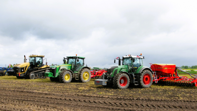 Trīs krāsaini traktori uz lauka ar piekabinātiem dažādiem agregātiem