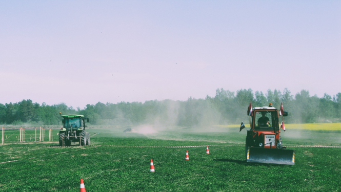 Divi krāsaini traktori brauc pa lauku. Sarkanais traktors brauc pretī, kamēr zaļais prom. Fonā meža ainava
