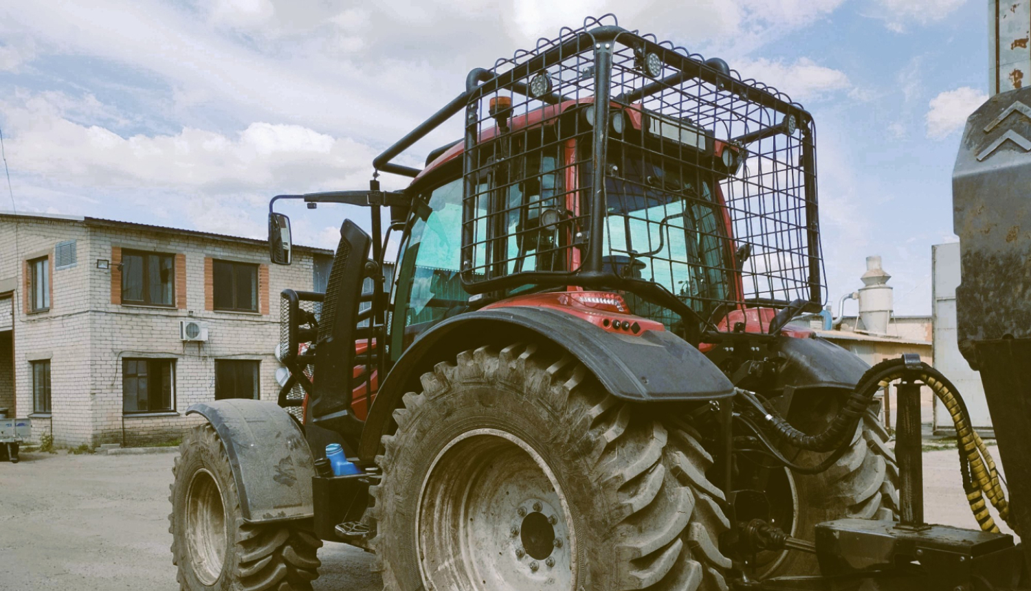 Sarkans traktors pieāķēts pie piekabes. Traktora kabīnei apkārt piestiprināts papildu drošības režģis