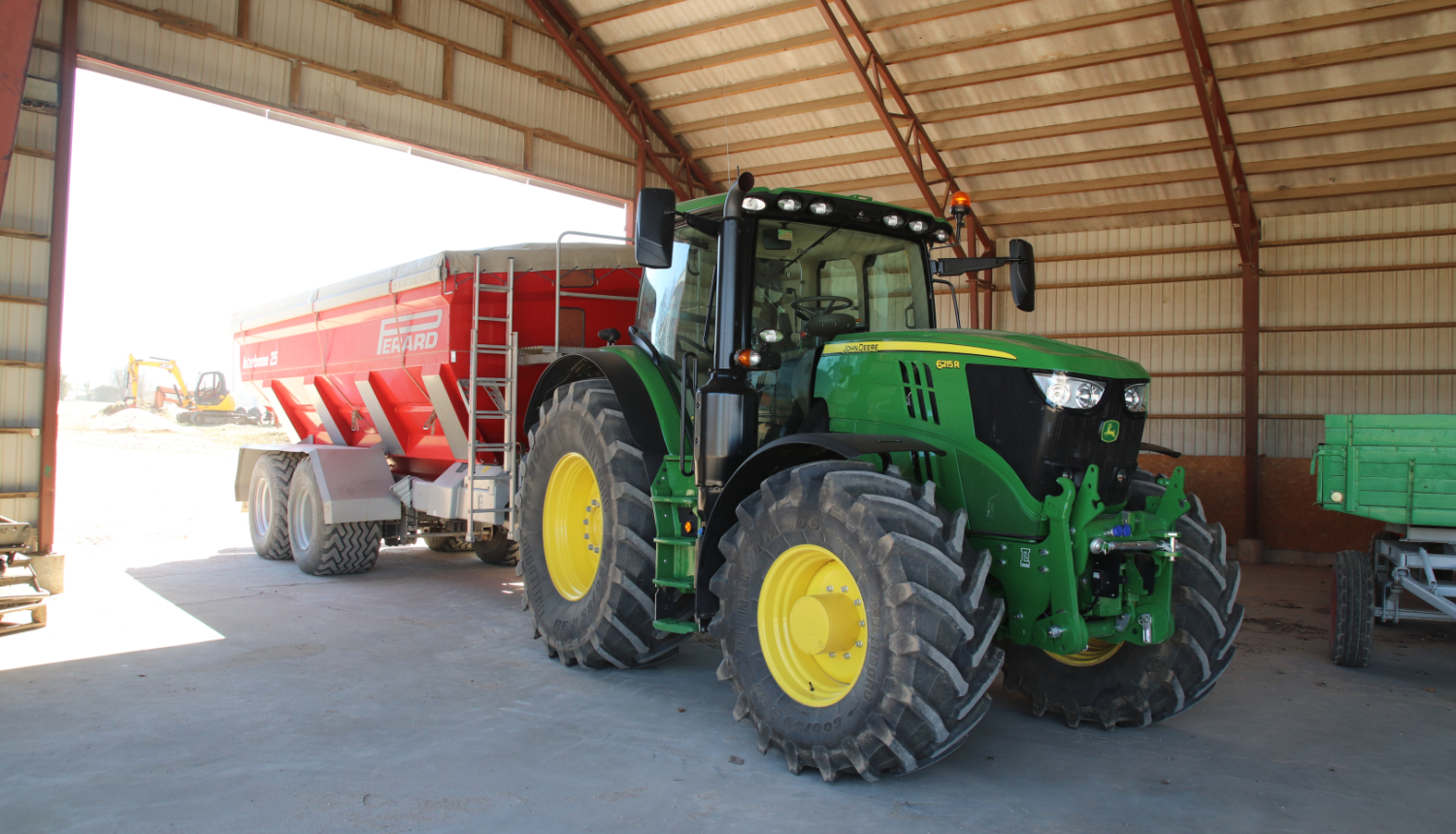 Zaļš traktors ar dzelteniem riteņiem un sarkanu piekabi iebraucis noliktavā. Attēls no traktora priekšas, skatā no noliktavas vidus