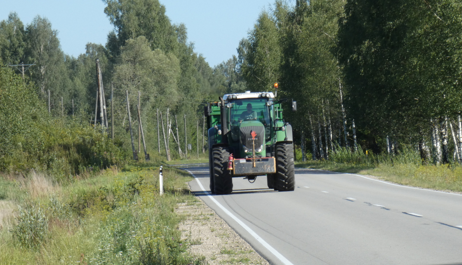 Zaļš traktors ar piekabi brauc pa asfaltētu ceļu. Fonā mežs