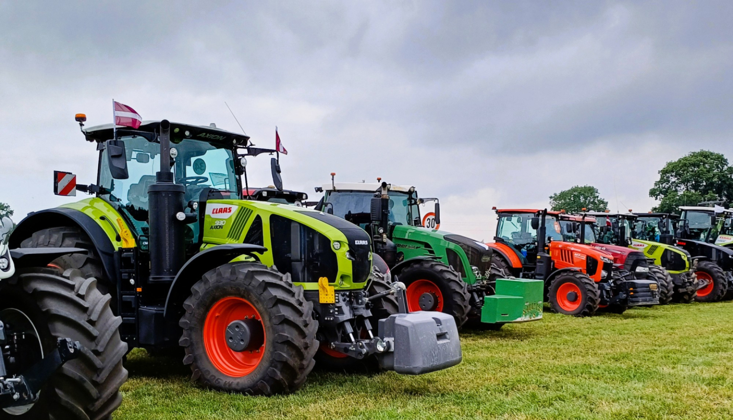 Krāsaini traktori vienā nevienlīdzīgā rindā uz zaļa lauka