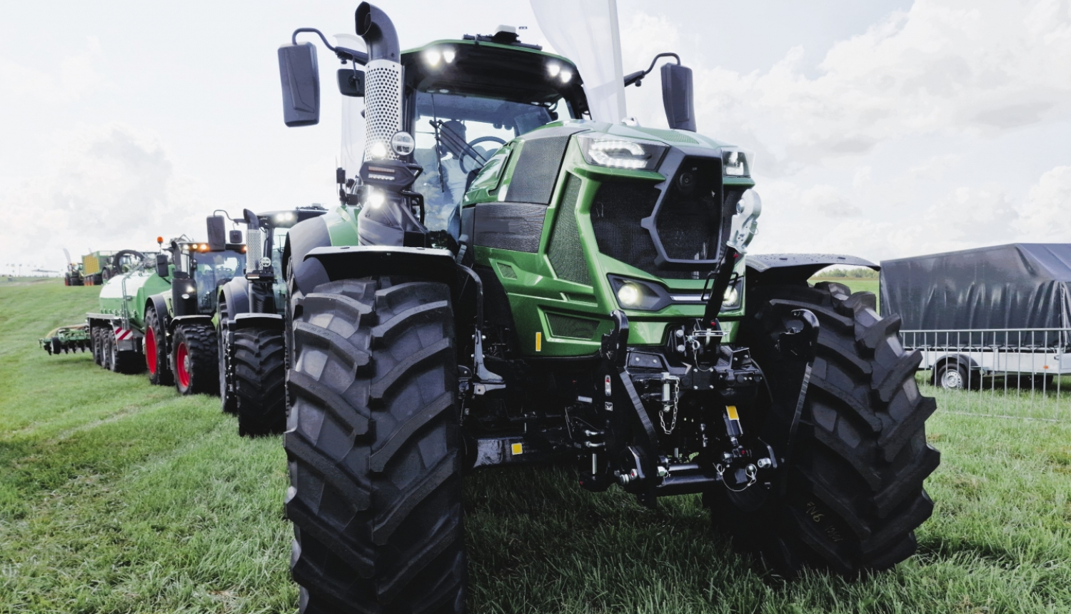 Traktoru kolonna pļavā, priekšā zaļš traktors ar lielām, melnām riepām