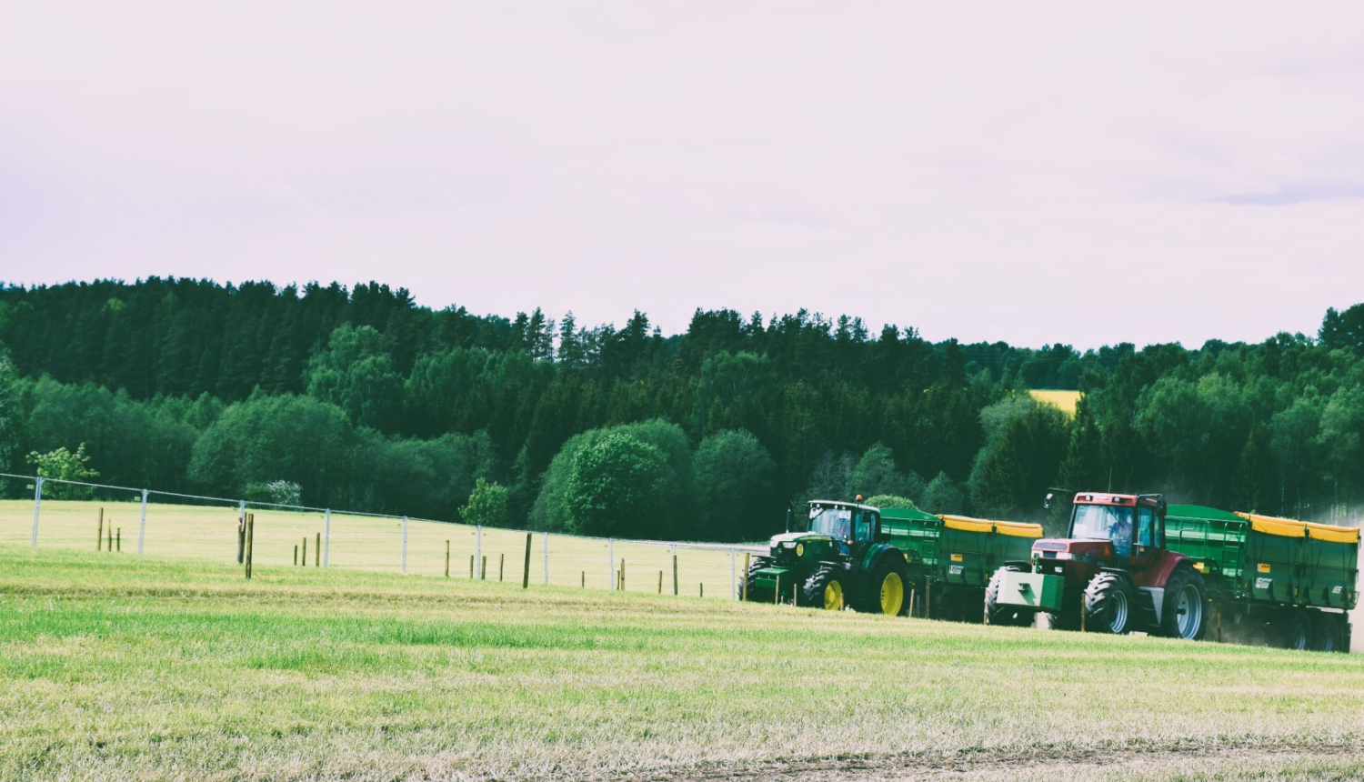 Divi krāsaini traktori brauc paralēli pret kalnu. Fonā meža ainava