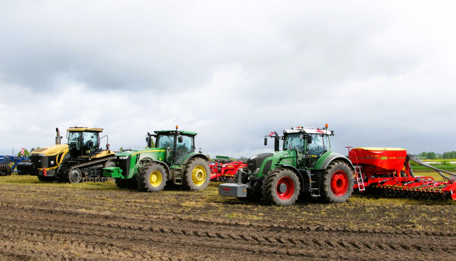 Trīs krāsaini traktori uz lauka ar piekabinātiem dažādiem agregātiem