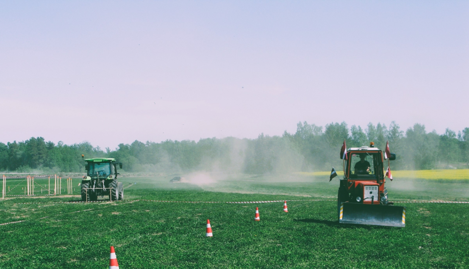 Divi krāsaini traktori brauc pa lauku. Sarkanais traktors brauc pretī, kamēr zaļais prom. Fonā meža ainava