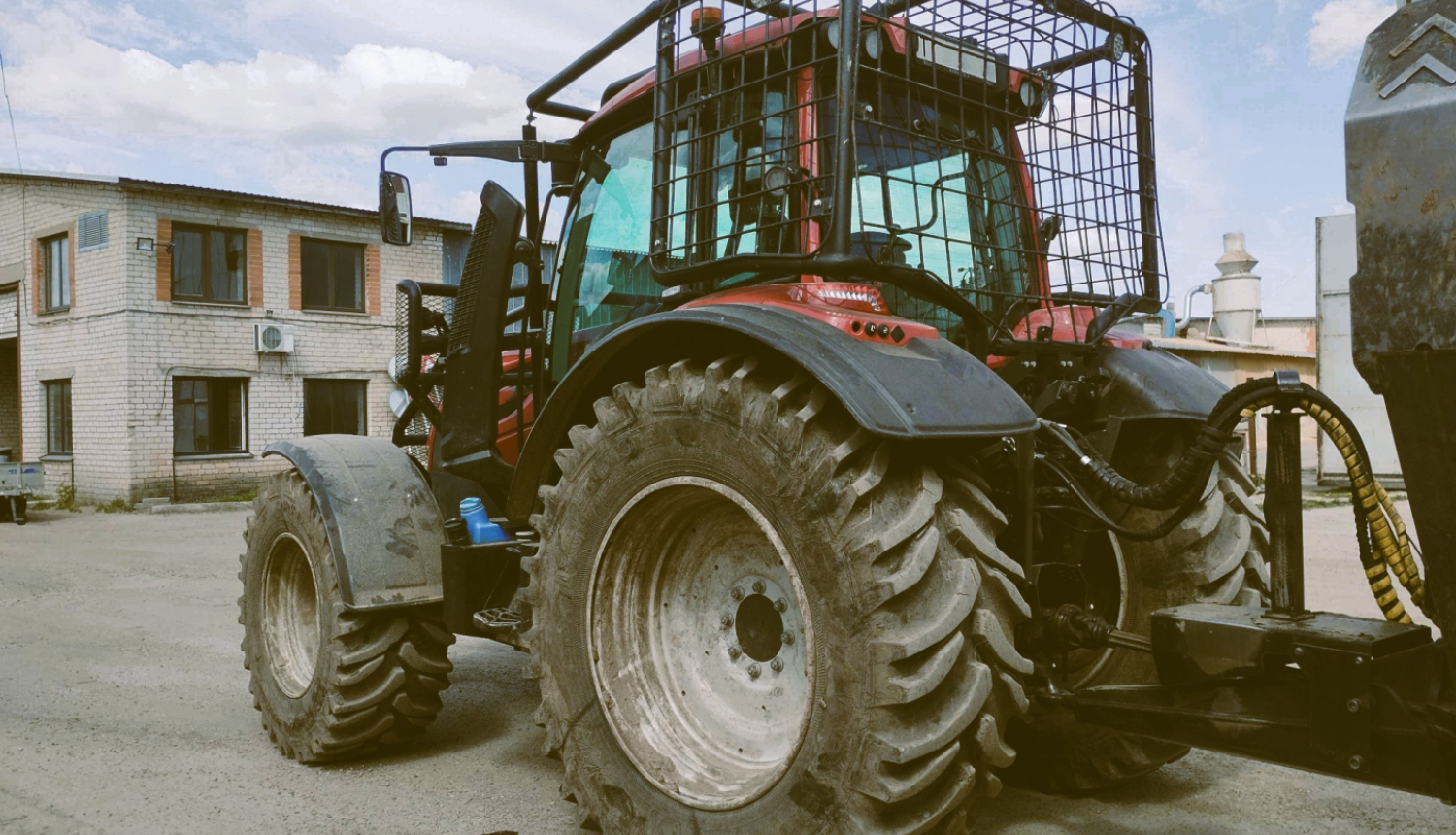 Sarkans traktors pieāķēts pie piekabes. Traktora kabīnei apkārt piestiprināts papildu drošības režģis