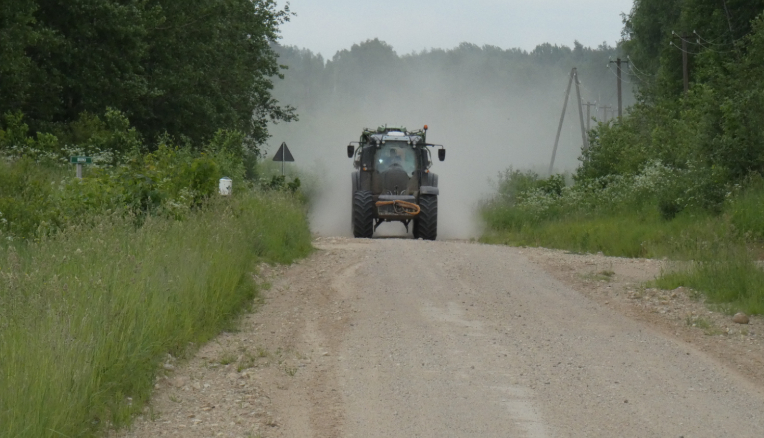 Tumšs traktors brauc pa zemes ceļu, veidojot pēc sevis lielu putekļu mākoni