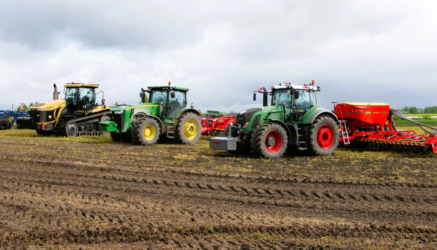Trīs krāsaini traktori uz lauka ar piekabinātiem dažādiem agregātiem