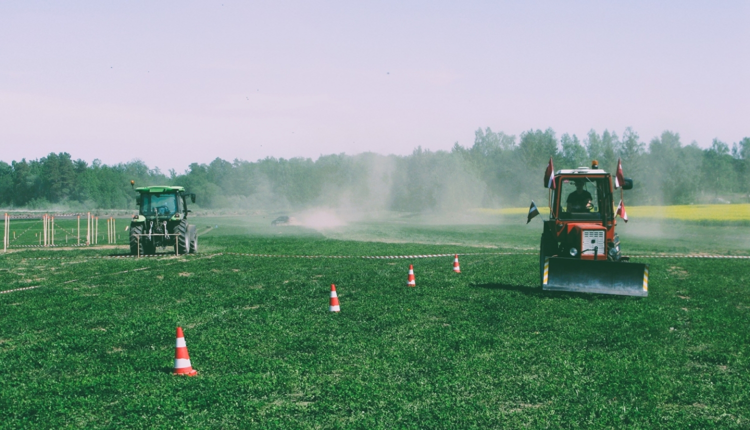 Divi krāsaini traktori brauc pa lauku. Sarkanais traktors brauc pretī, kamēr zaļais prom. Fonā meža ainava